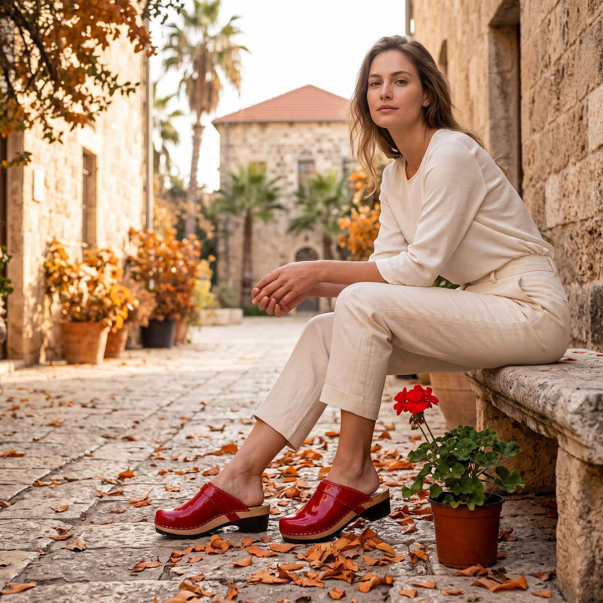 דוגמנית בצילום חוץ ברחוב עתיק ספסל אבן עציץ עם פרח אדום, לובשת חולצת טריקו לבנה, מכנס בד פשתן צמוד וקבקבי עץ בצבע אדום לק בוהק מיוחדות עם סוליה שקטה מתחת לבסיס העץ לכפכף נוח ושקט יותר בייבוא מיוחד משבדיה של ג׳ילון סטודיו לישראל