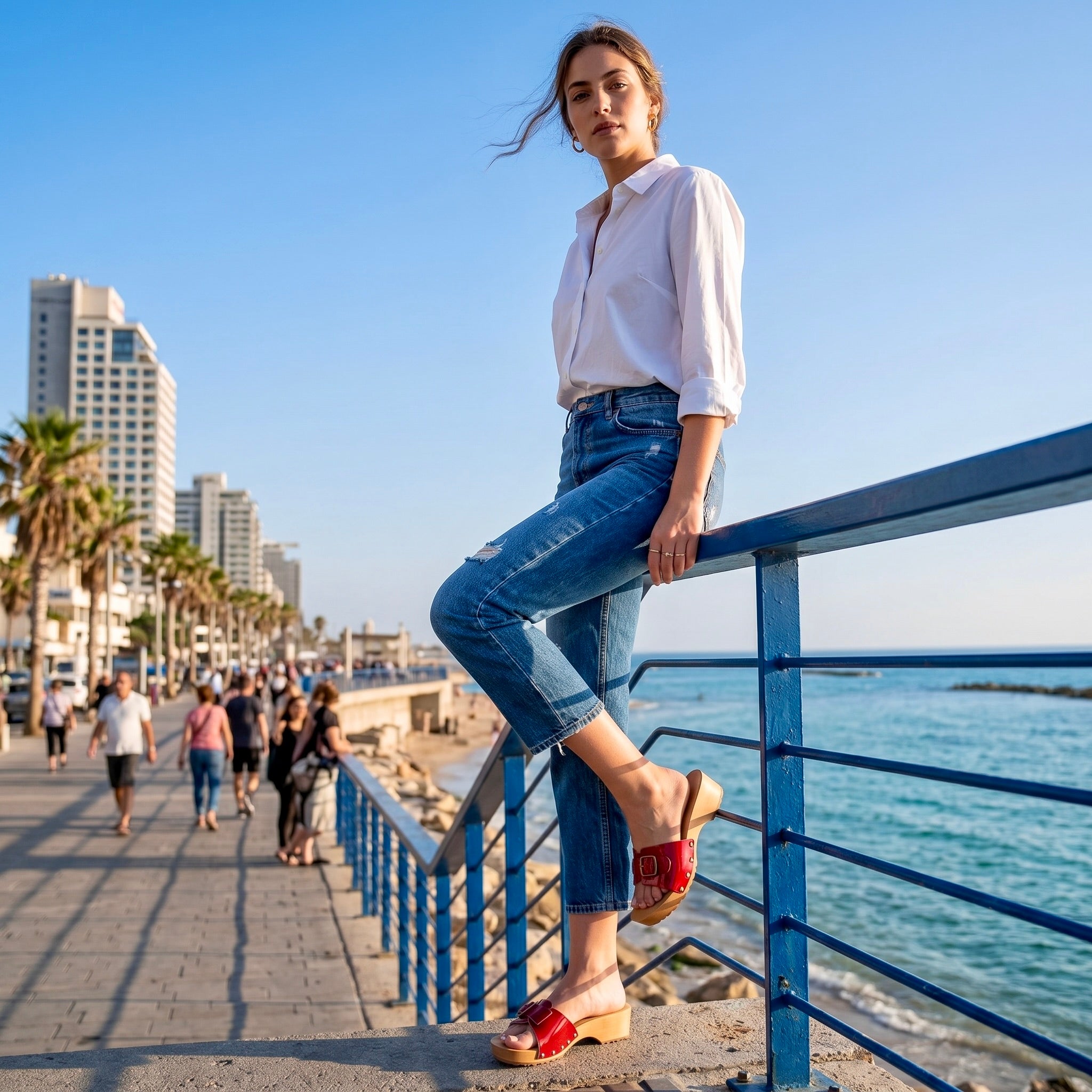 "פרופיל של דוגמנית בטיילת חוף אורבנית, לבושה בג'ינס וחולצה לבנה, מניחה רגל על מעקה. היא נועלת קבקבי עץ אופנתיים לנשים בעלי רצועת עור אדומה מבריקה ואבזם זהב, המשלבים סטייל קיצי ונוחות מול הים