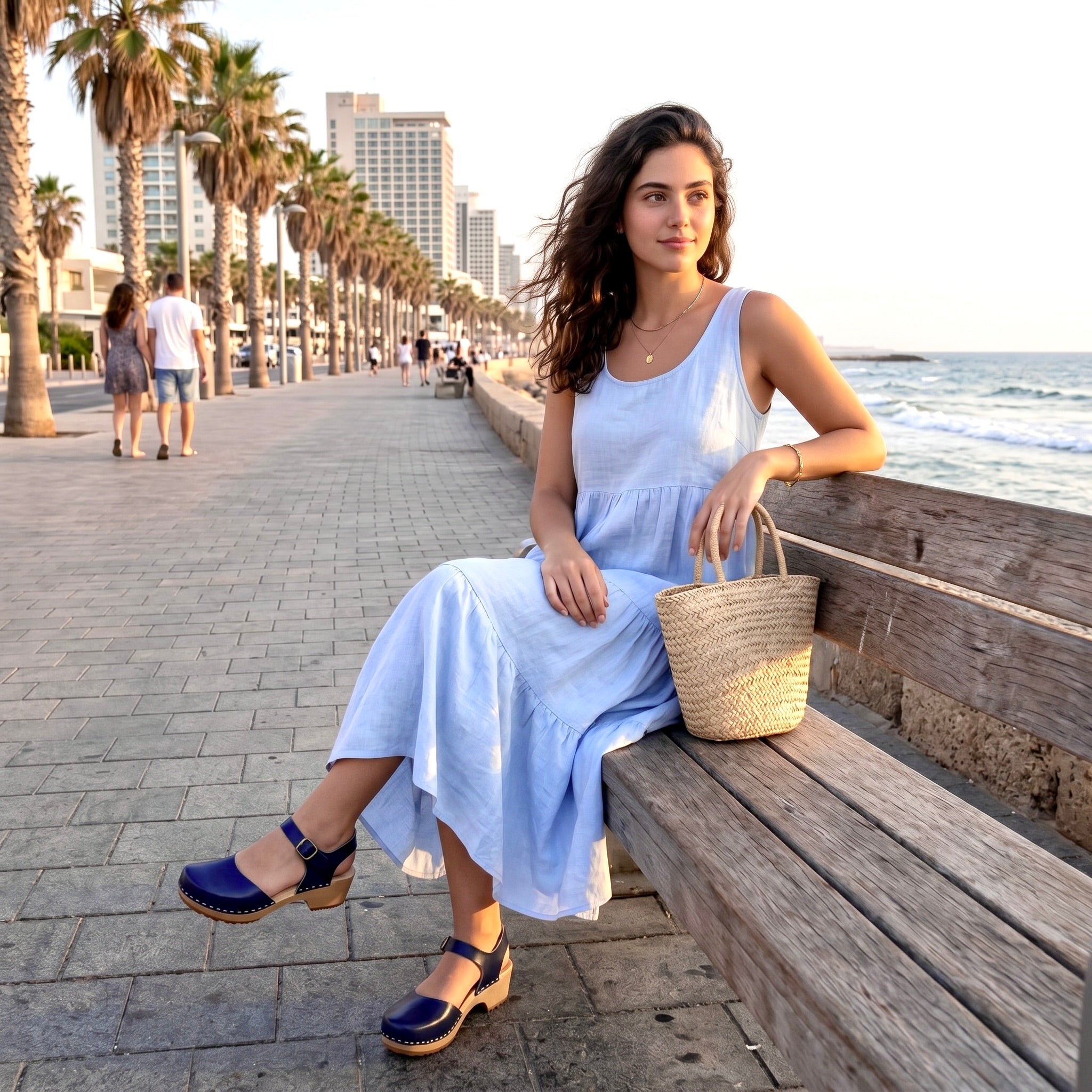בחורה נועלת סנדל כחול נייבי, לבושה שמלה קיצית בגוון תכלת עם תיק קש יושבת על ספסל ציבורי ליד החוף בטיילת תל אביב
