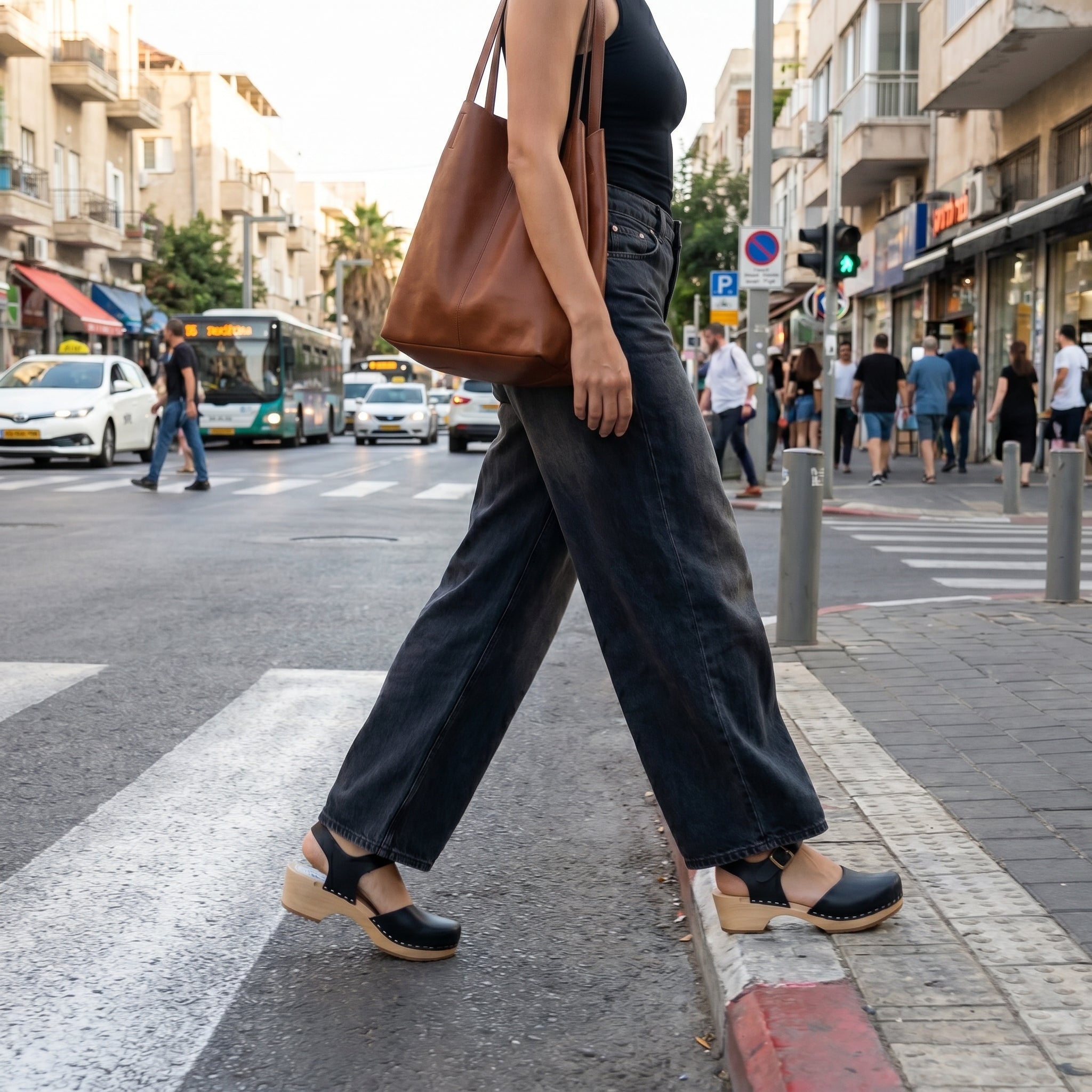 מבט מהצד על דוגמנית פוסעת על מעבר חצייה עירוני, לובשת מכנסיים רחבים כהים וגופיה שחורה עם תיק עור חום, ונועלת סנדלי קבקב אמה (Emma) מעור שחור עם עקב בלוק נמוך וסוליית עץ