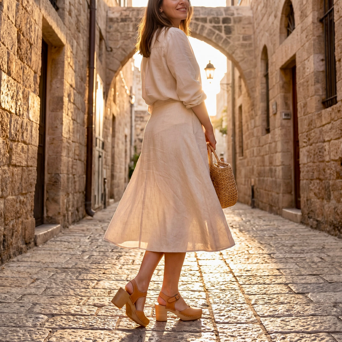 תקריב בגובה הרצפה של רגלי אישה עומדות על מדרחוב מרוצף אבן בסמטה עתיקה בשעת שקיעה, לבושה בחצאית בהירה ונועלת סנדלי קבקב בגוון דבש עם עקב גבוה. מקולקציית קבקבי-העץ השוודיים של ג׳ילון-סטודיו