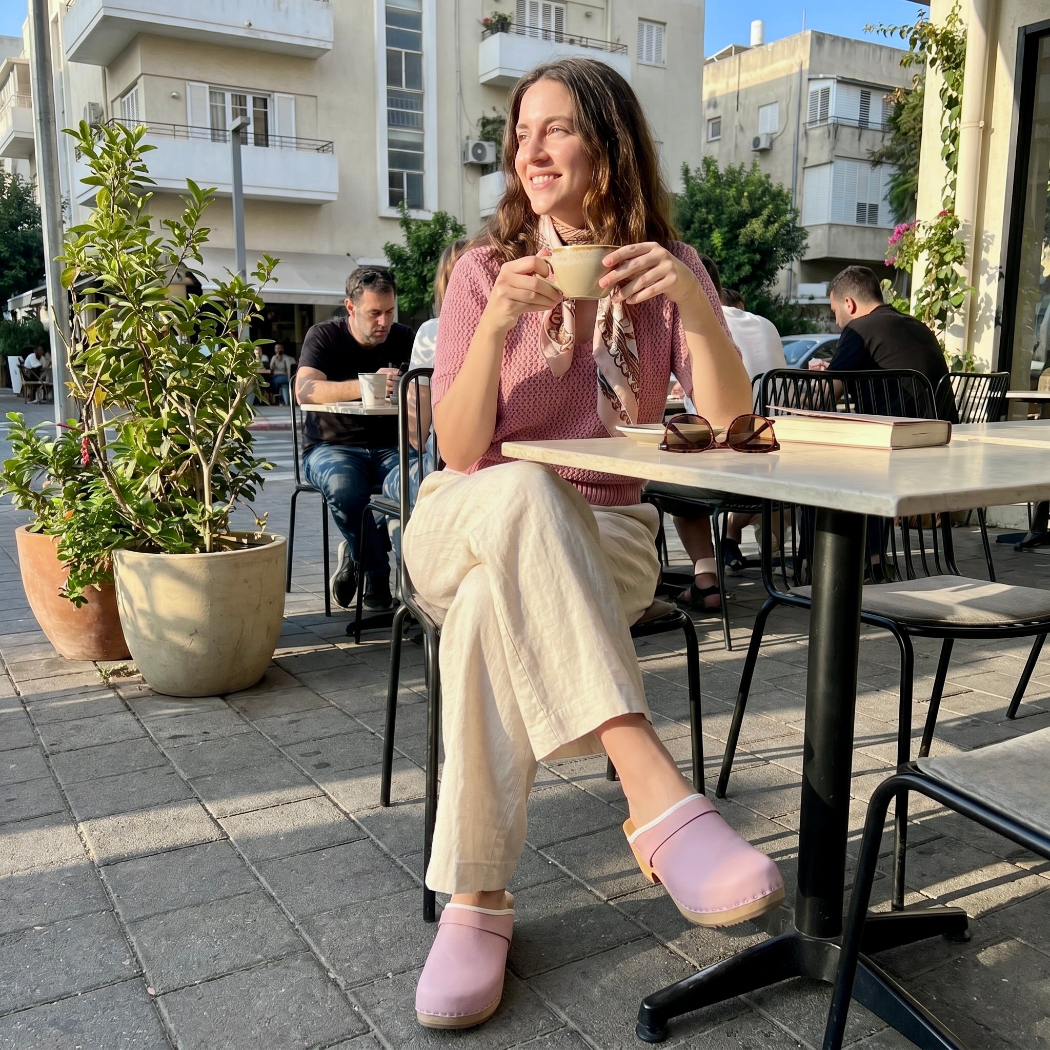 אישה יושבת על כיסא עץ ליד שולחן בחלל בית קפה חמים, לבושה בז'קט ומכנסיים בהירים ונועלת קבקבים בגוון ורוד פודרה.