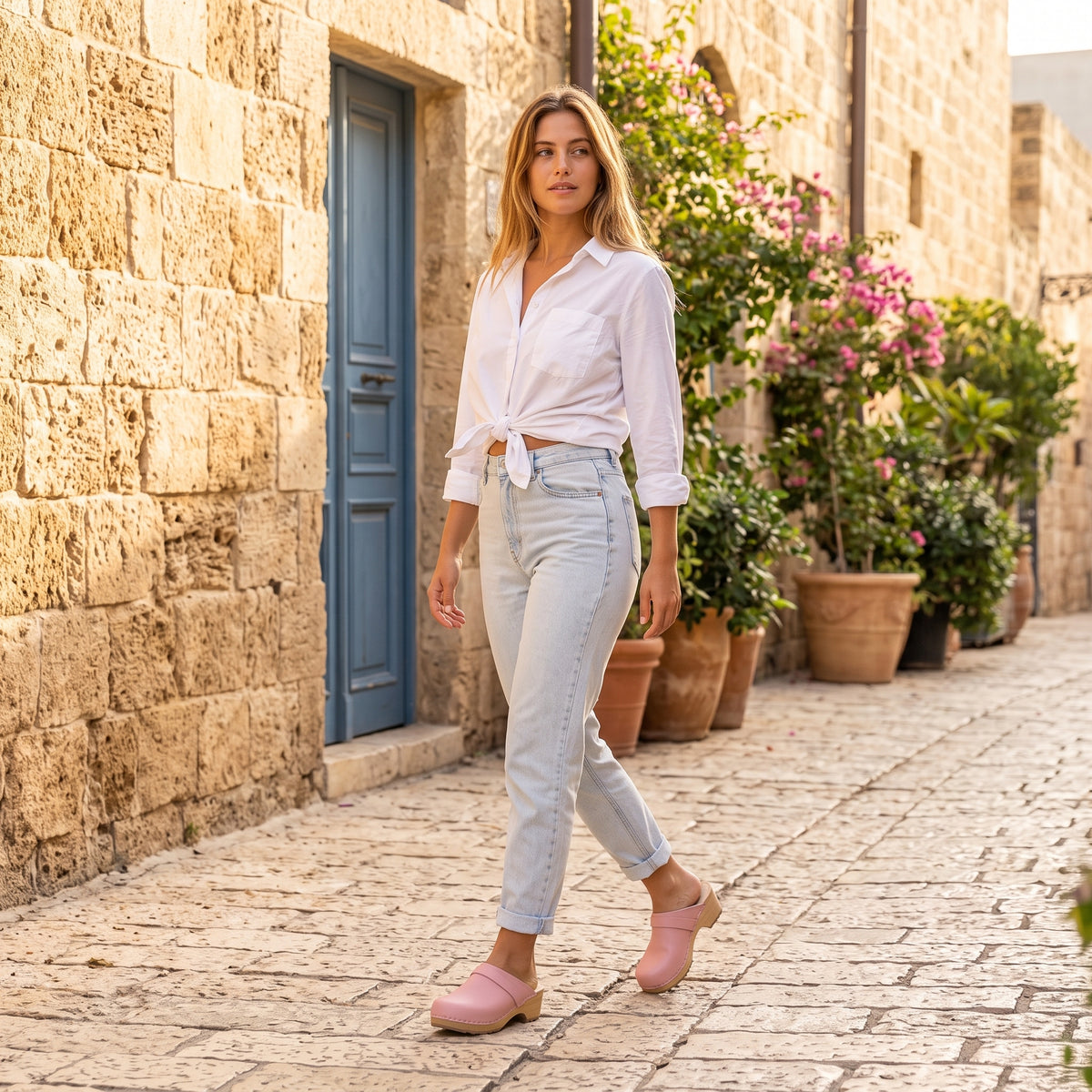 דוגמנית במכנס ג׳ינס ארוך וחולצה לבנה בסמטת אבן ירושלמית, נועלת קבקבי-עץ שוודיים של ג׳ילון-סטודיו בצילום הליכה טבעית