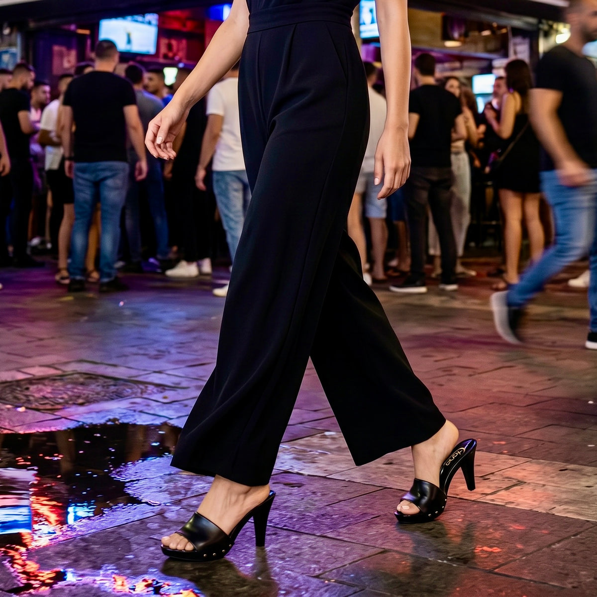 צילום ממוקד מהמותניים ומטה בדוגמנית הפוסעת בשעת ערב על מדרכה רטובה ומשקפת אורות ברחוב עירוני הומה אדם, כשהיא לובשת אוברול שחור ארוך ונועלת קבקבי עץ לנשים מעור שחור חלק.