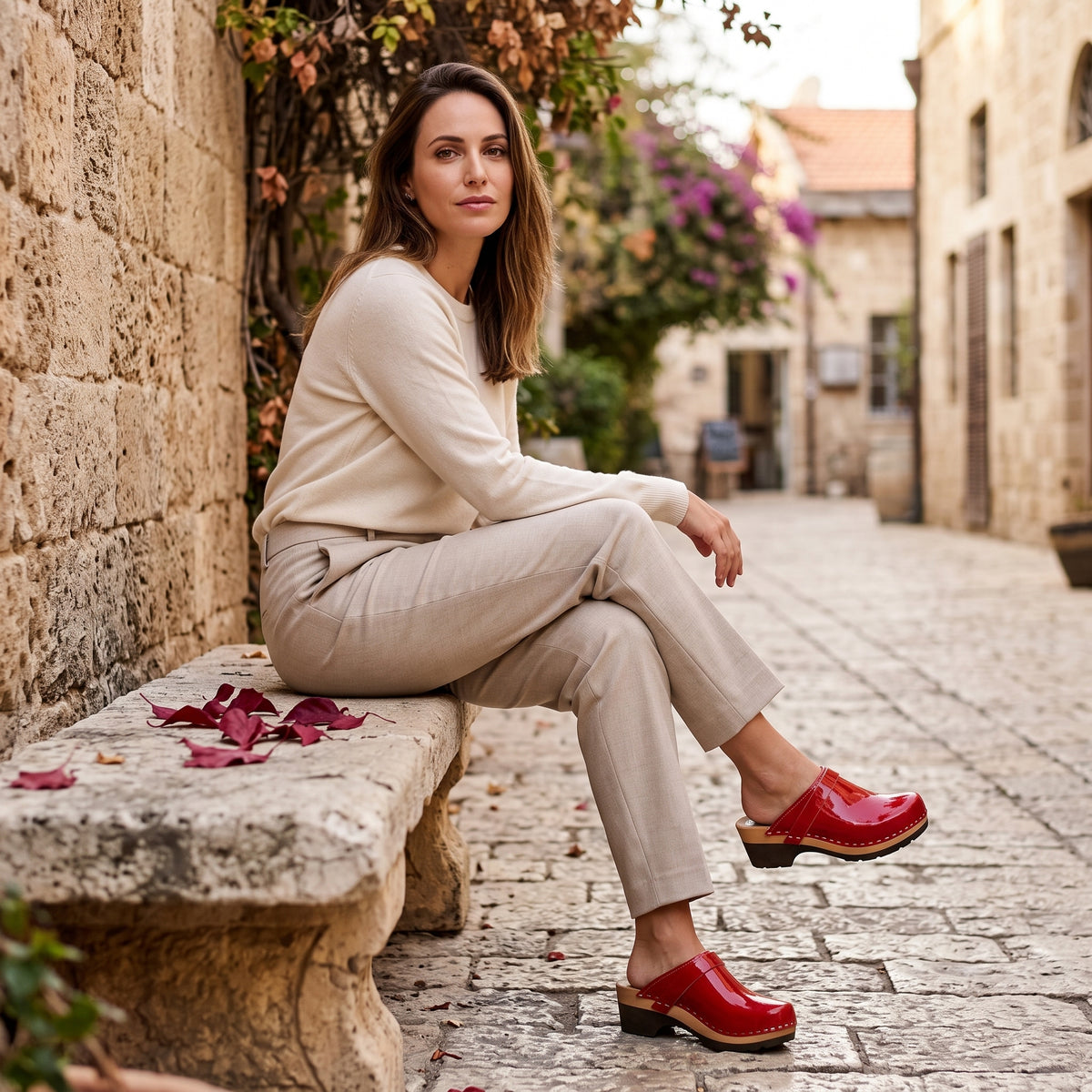דוגמנית בצילום חוץ ברחוב עתיק ספסל אבן עלי שלכת אדומים, לובשת חולצת טריקו לבנה, מכנס בד פשתן צמוד וקבקבי עץ בצבע אדום לק בוהק מיוחדות עם סוליה שקטה מתחת לבסיס העץ לכפכף נוח ושקט יותר בייבוא מיוחד משבדיה של ג׳ילון סטודיו לישראל