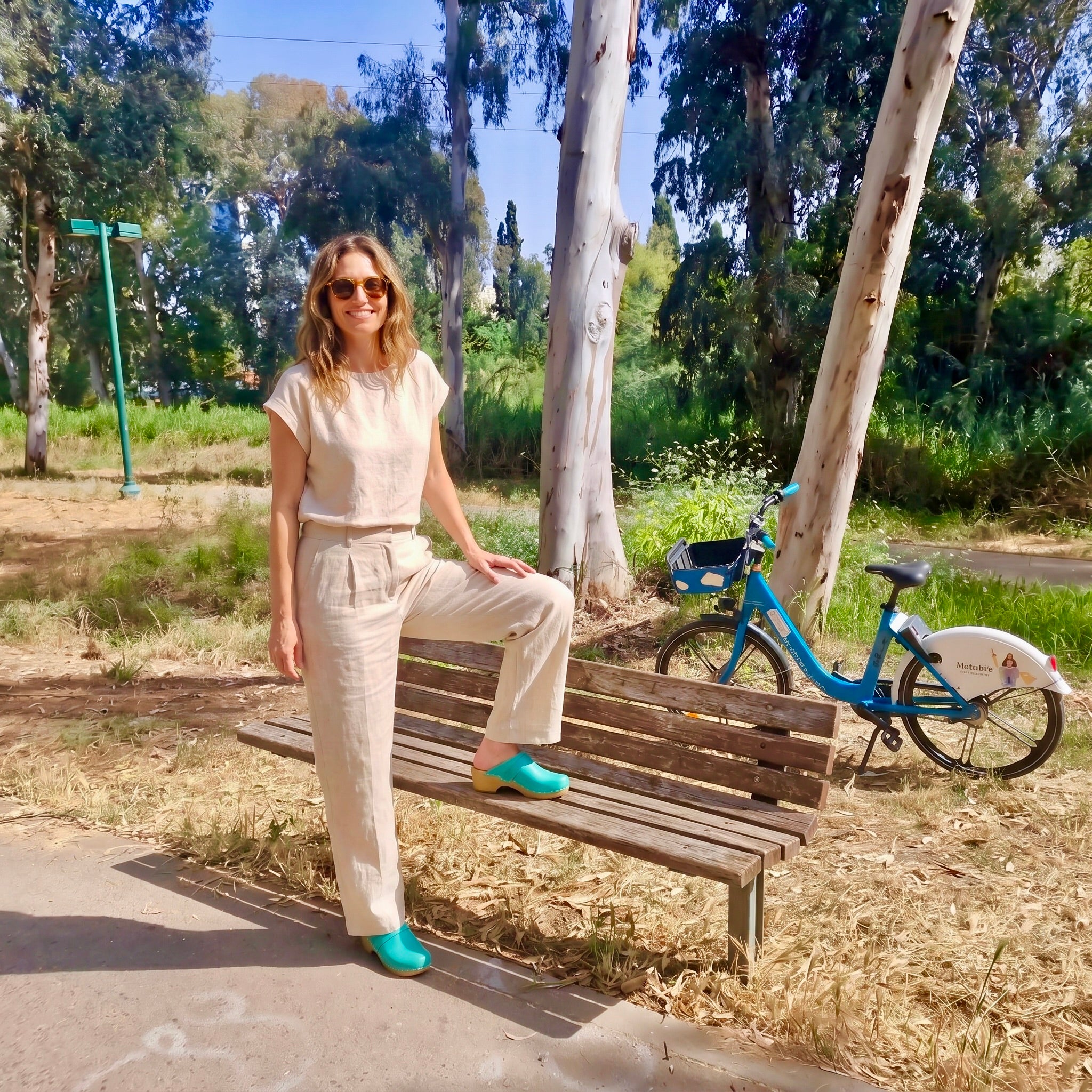 אשה משעינה רגל אחת על ספסל בפארק הירקון, קבקב ירוק פיסטוק על רקע אופניים וחורש ג׳ילון סטודיו מקולקציית קבקבי-העץ השוודיים של ג׳ילון סטודיו
