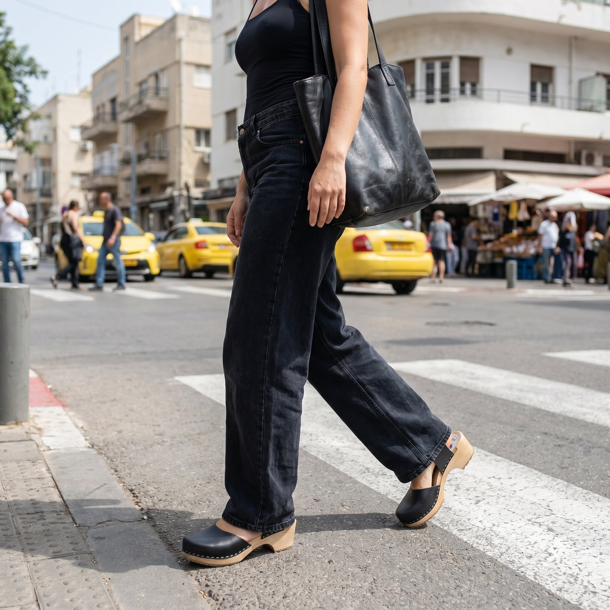 דוגמנית בתנועת הליכה על מעבר חצייה ברחוב עירוני, לובשת ג'ינס שחור רחב, גופיה שחורה ותיק כתף מעור שחור, נועלת סנדלי קבקב אמה (Emma) עם עקב עץ נמוך ויציב