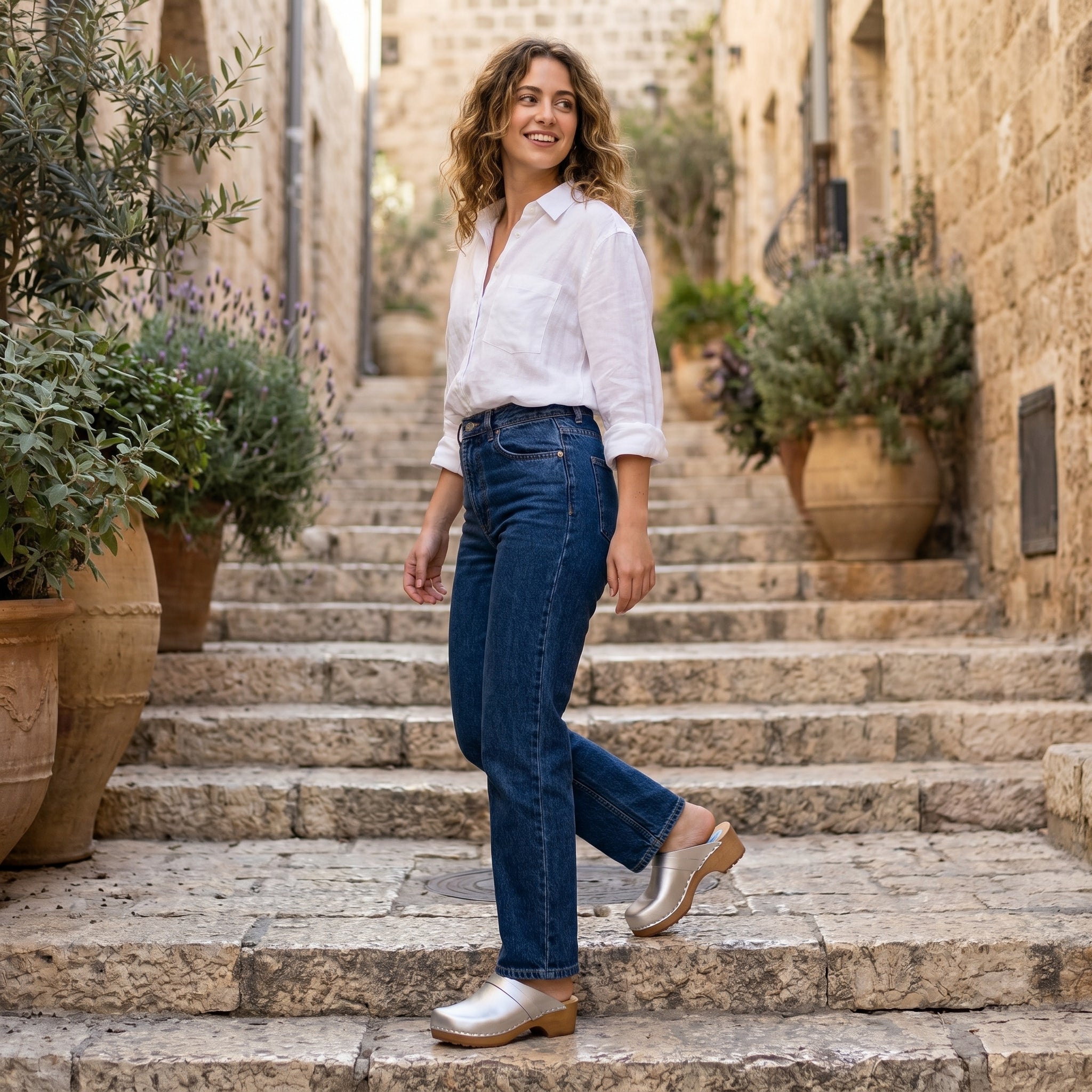 אישה יורדת במדרגות אבן חיצוניות, לבושה בחולצה מכופתרת לבנה, מכנסי ג'ינס כחולים וקבקבים לבנים.