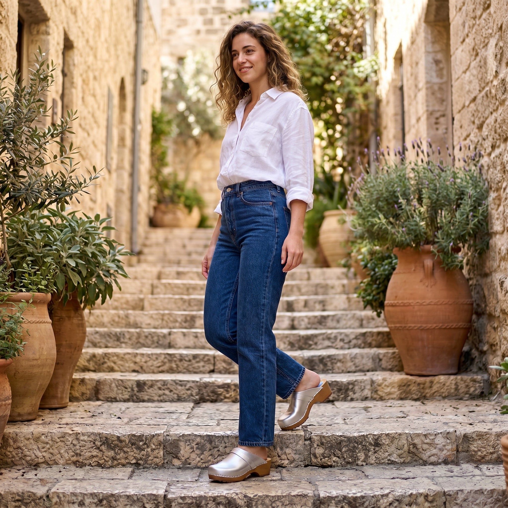 אישה עומדת על מדרגות אבן חיצוניות לצד כדי חרס גדולים, לבושה בחולצה לבנה, ג'ינס כחול וקבקבים לבנים.