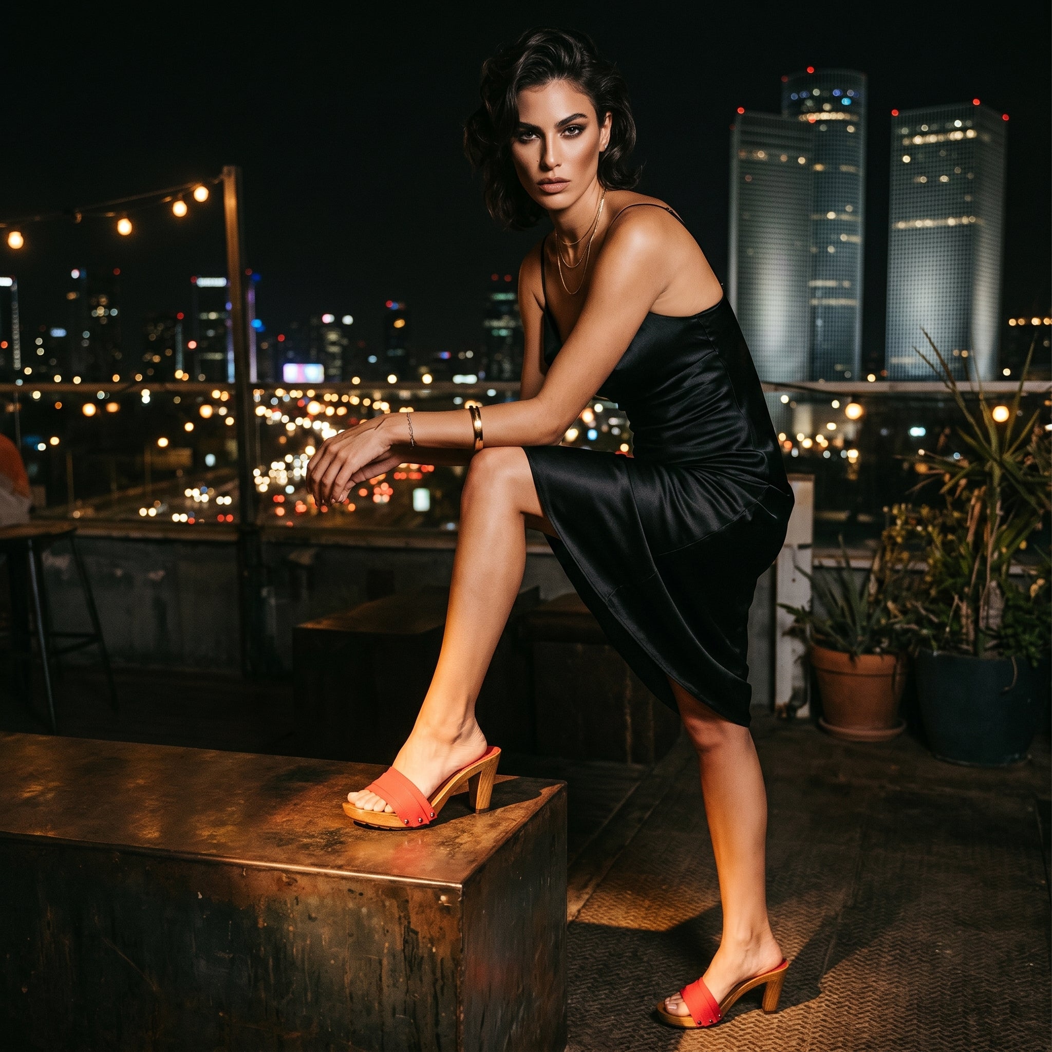 צילום אופנה לילי על גג (Rooftop) מעוצב מול קו הרקיע המואר של העיר ותאורת מגדלים. הדוגמנית נשענת על ארגז מתכת, לובשת שמלת סאטן שחורה עם כתפיות דקות ונועלת את קבקבי עץ הזמש האדומים מסדרת סופיה קומפורט.