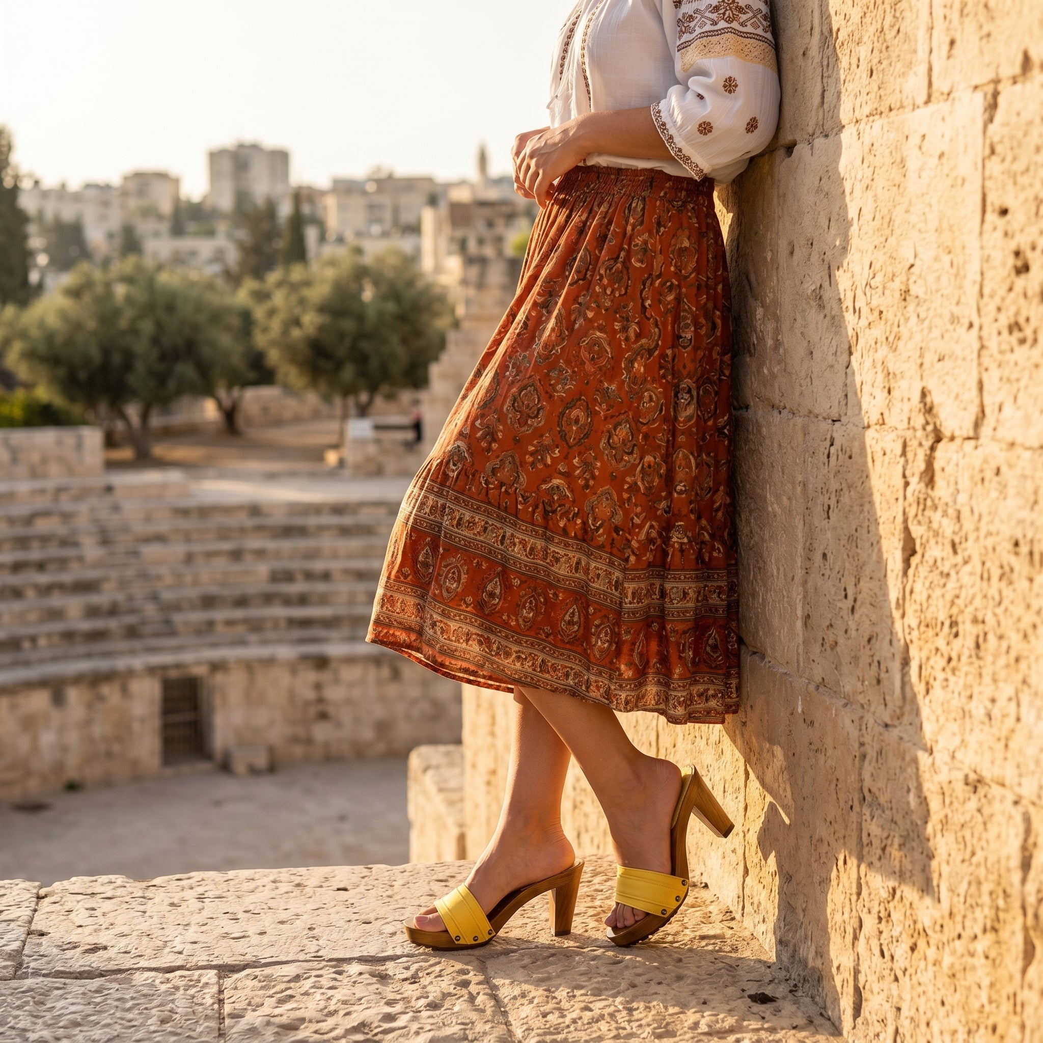 קלוז-אפ מהמותניים ומטה של דוגמנית נשענת על קיר אבן עתיק ומחוספס בשעת שקיעה. היא לובשת חצאית בוהו-שיק ארוכה ומודפסת בגווני חום ואדמה, ונועלת את קבקבי עץ הזמש הצהובים דגם סופיה קומפורט המציגים את עקב הבלוק היציב בגובה 8 ס"מ.