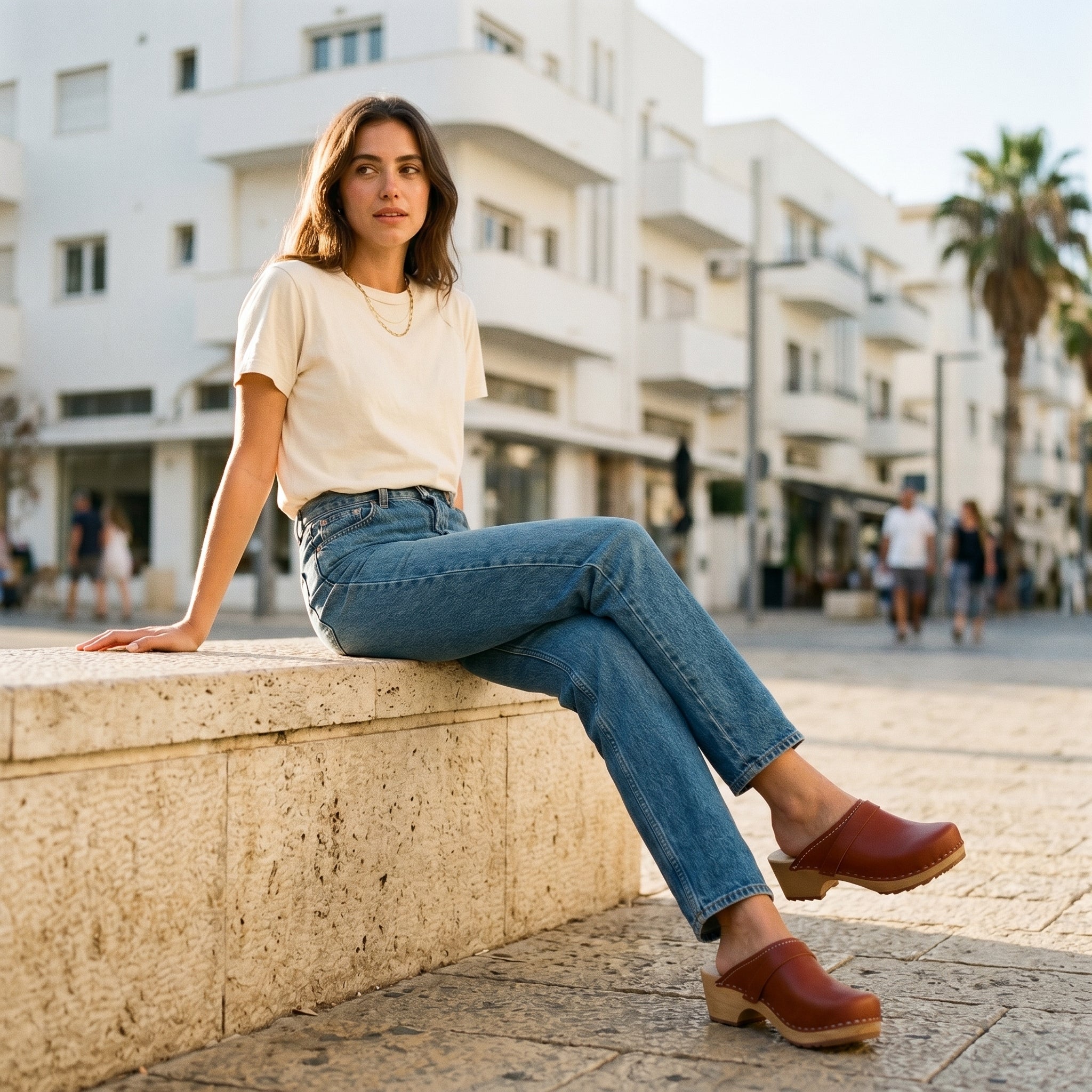 דוגמנית יושבת על חומת אבן ברחוב תל אביבי, מציגה את קבקב אנה קרמל בשילוב ג'ינס וטי-שירט לבנה.