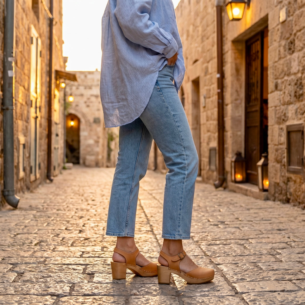 תקריב מהצד של רגלי אישה עומדת בסמטת אבן עתיקה, לבושה בג'ינס כחול ישר וחולצה תכולה מכופתרת אוברסייז, ונועלת סנדלי קבקב בגוון דבש עם עקב גבוה. מקולקציית קבקבי-העץ השוודיים  של ג׳ילון-סטודיו